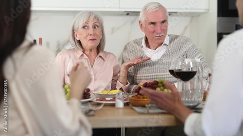 Mature spouses sitting at table, enjoying food and wine, chatting talking laughing with adult son and his girlfriend. In homely atmosphere male and female pensioner celebrate anniversary with guests
