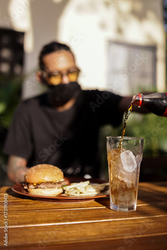 A person pours cold soda into a glass filled with ice, with a plate of burger and fries on the table. The meal looks fresh and casual, perfect for lunch, snack, or fast food moments.