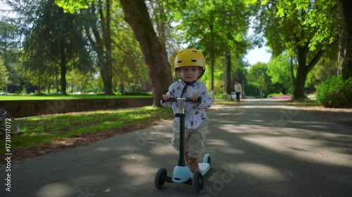Wallpaper Mural Small toddler wearing yellow helmet riding scooter along tree-lined park path, moving with curiosity and energy while exploring outdoor environment Torontodigital.ca