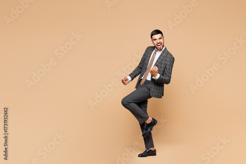 Full body young happy successful employee business man corporate lawyer wear classic formal grey suit shirt tie work in office do winner gesture raise up leg isolated on plain beige background studio.