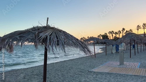 Scenic beach view with straw parasols and calm sea waves at sunset. The sandy shore and rustic umbrellas create a relaxing summer holiday atmosphere, perfect for themes of travel, vacation, leisure