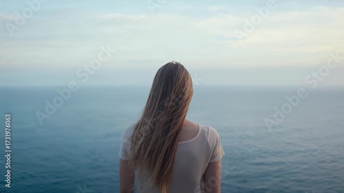 Feeling gentle breeze picking up, lone woman lifting hair while scanning ocean horizon from cliff