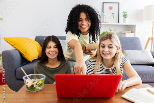 Wallpaper Mural Three cheerful multi-ethnic young women using laptop while eating takeaway food in living room. Female friendship and youth concept Torontodigital.ca