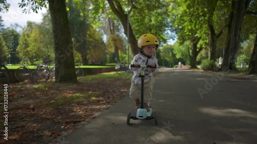 Wallpaper Mural Toddler wearing yellow helmet pushing scooter forward on shaded park path, full of energy and determination while enjoying outdoor playtime activity Torontodigital.ca