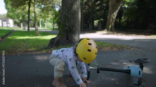 Toddler with yellow helmet falling forward off scooter on paved park path, child stumbling during outdoor play and landing on ground in sudden accident