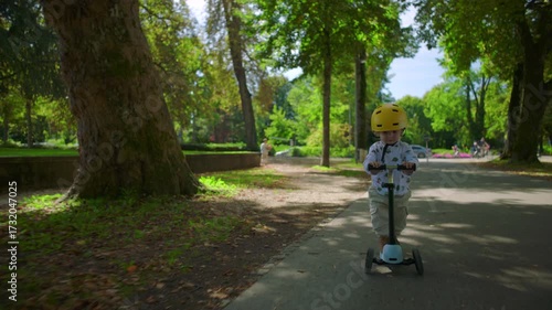 Wallpaper Mural Toddler with yellow helmet riding scooter down shaded park path, small child concentrating while practicing balance and movement outdoors Torontodigital.ca