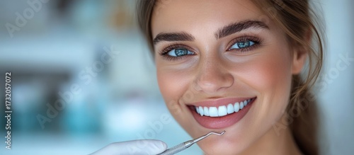 Smiling woman in dental setting. Bright, white teeth, blue eyes, dental tools visible. Close-up portrait