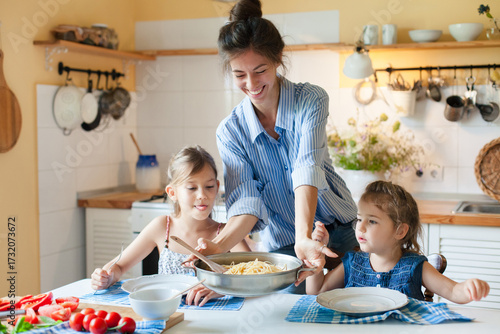 Happy mother serving pasta for daughters at home kitchen. Cozy family lunch, parenthood, comfort food, healthy eating for kids concept. Homemade Italian cuisine, mac and cheese. Authentic lifestyle
