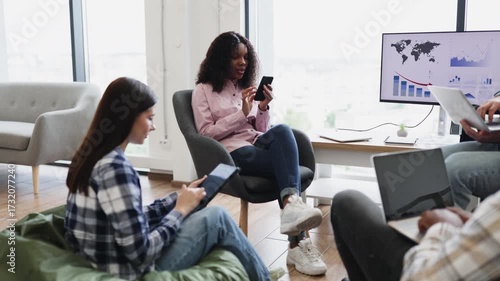 Two female colleagues, one brunette and one curly-haired, using smartphones while sitting in modern office space during informal discussion. Scene conveys teamwork, communication, technology use