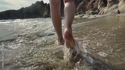 A close-up of bare feet walking along a wet sandy beach near the shoreline, with gentle waves washing over nearby rocks. The sunlight casts a soft shadow, enhancing the natural textures of sand, water