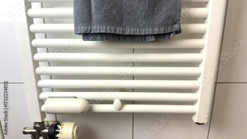 Bathroom radiator heater with gray towel, white wall mounted heating and towel drying system, clean textile and thermostat, heating equipment and modern bathroom interior.