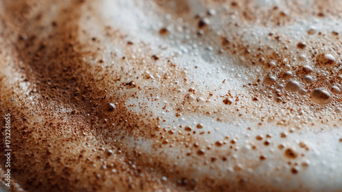 The image presents a close up view of a beverage surface, primarily composed of white, frothy foam. Dark, powdery substance is scattered across the foam, creating a textured appearance