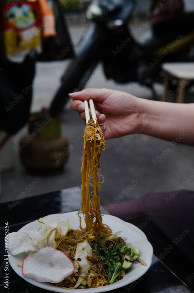 Φωτογραφία A person's hand using chopsticks to lift a portion of Indonesian fried rice vermicelli (bihun goreng) from a plate at an outdoor street food stall