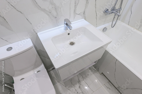 Clean, modern bathroom featuring white fixtures: a toilet, a sink with a chrome faucet, and a bathtub. Marble-patterned tiles on the floor and walls add a touch of elegance