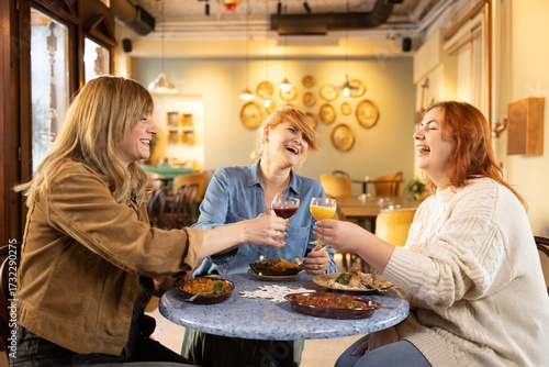 Three female friends enjoying drinks and food, laughing together at restaurant table