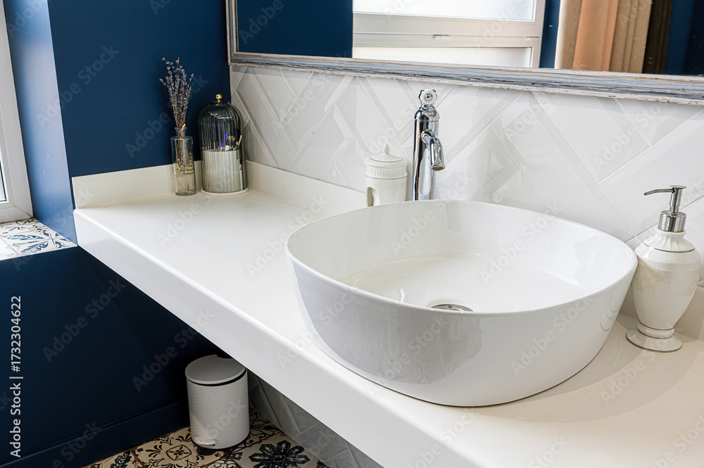 Fototapeta premium Clean bathroom featuring a round white sink, white countertop, blue wall, herringbone tile backsplash, and decorative accessories
