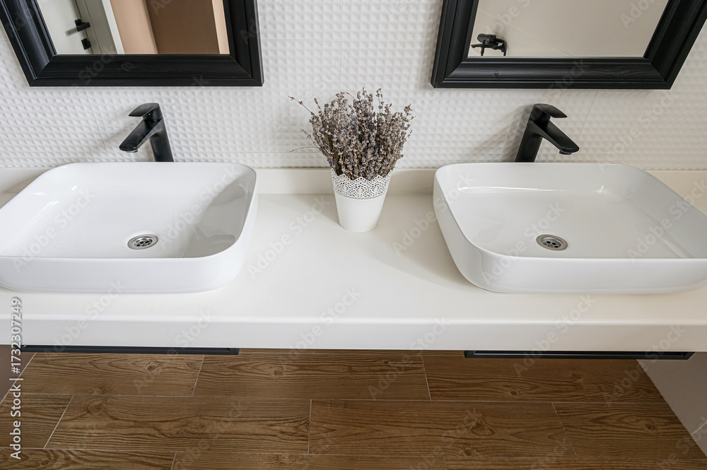 Fototapeta premium A modern bathroom with two square, white sinks, black faucets, black-framed mirrors, a white countertop, and a pot of dried flowers