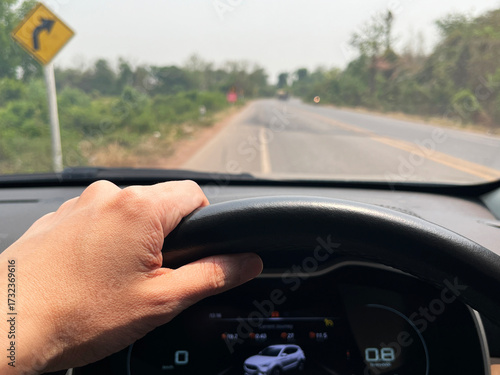 driving on a rural road with a visible curve warning sign, showing a close-up of a left hand holding the steering wheel inside a car, focusing on safe travel and road journey atmosphere dashboard view