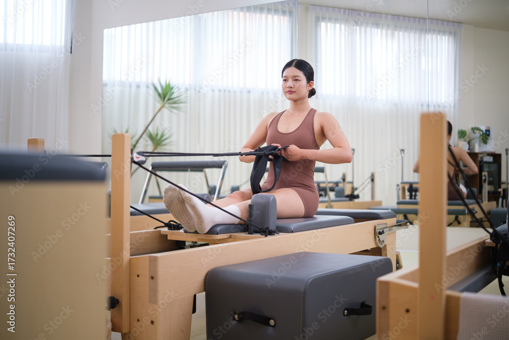 Fototapeta premium Woman performs a seated Pilates workout using resistance bands on a reformer machine in a modern studio.
