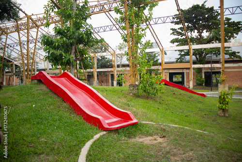 A bright red slide stands on a grassy hill in an outdoor playground beneath a metal frame structure.