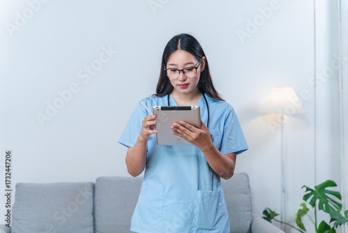 Young female healthcare professional in scrubs using tablet device in modern home environment while looking focused on the screen