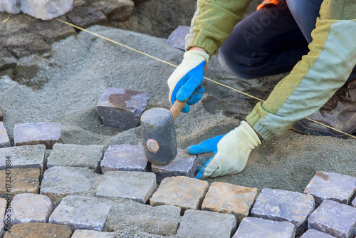 Laborer carefully places cobblestones levels sand for new pathway at construction site on work day.