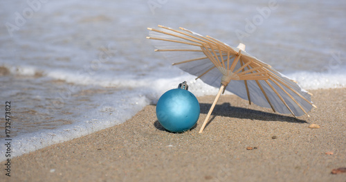 Blue Christmas bauble under beach umbrella on golden sand as sea wave approaches, creative minimalistic festive background for tropical holiday season and design with copy space