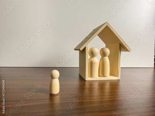 A small wooden house shelters two peg figures, while a third stands alone outside on a dark wooden surface with a white background.