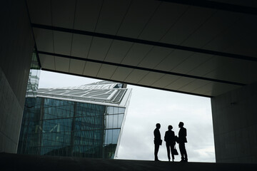 Three middle aged business professionals standing in modern urban setting, silhouettes engaged in conversation near glass office building, diverse group interacting outdoors