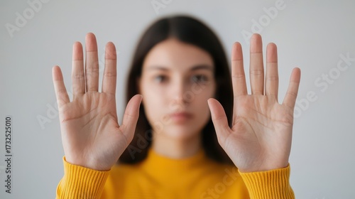 A young woman in a yellow sweater holds her hands up, conveying a message of stop or pause, with a neutral background emphasizing her expression.