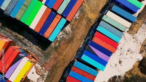 Aerial top-down view of a shipping container yard. Rows of brightly colored containers — red, blue, green, yellow — create a vibrant geometric pattern on dusty ground