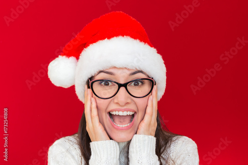 Young pretty caucasian brunette woman, wearing glasses, Santa hat and knitted white sweater looking at camera smiling in surprise on a red background