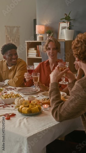 Vertical shot of red-haired young man proposing toast and clinking glasses with cheerful friends while celebrating thanksgiving at festive table