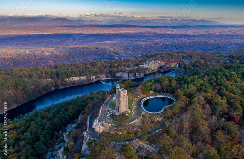 Aerial view of the stark stone tower of the Mohonk Preserve standing tall against the vibrant autumnal foliage and the tranquil waters of the lake, New York, United States.