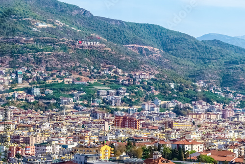 View of the Alanya hillside in Turkey with the I Love Alanya sign visible, surrounded by residential buildings. The background showcases the green mountains and the urban landscape of the Turkish city