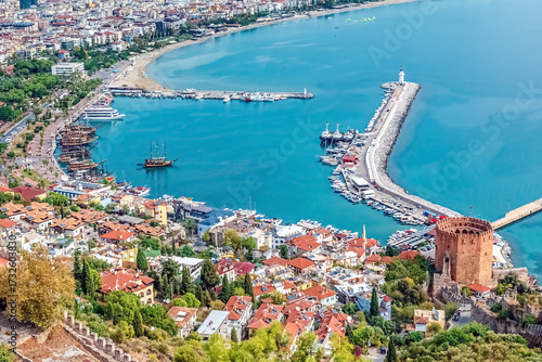 Aerial view of Alanya Harbor in Turkey, featuring the Red Tower and historic walls of Alanya Castle. The photo, taken from Alanya Castle, showcases the marina and the lighthouse