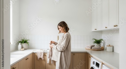 A pregnant laundry woman gently folds baby clothes in a bright, modern, minimalist laundry room with white tiles and wooden cabinetry, conveying care and anticipation.