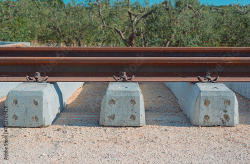 Close-up of a rusted railway rail, metal fasteners, and new concrete sleepers set against a textured gravel foreground and blurred olive tree background.