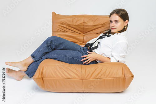 Studio shot: a stylish young woman in blue jeans and a white shirt poses lying crosswise in a chair against a white background.