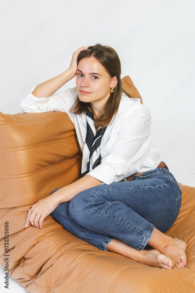Fototapeta premium Studio shot: a stylish young woman in blue jeans and a white shirt posing while sitting in a chair against a white background