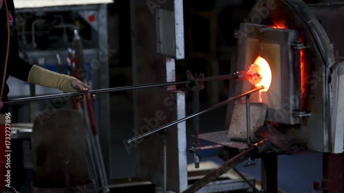 Close-up glassblowing process tube tool with molten glass shaped inside refractory furnace in industrial craft workshop. Handmade blowing glassware design production manufacture Europe