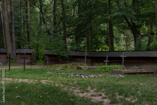 This peaceful image depicts a quaint rural cemetery featuring several weathered wooden crosses standing in a lush green grassy field. Surrounded by dense forest trees and small wooden structures 