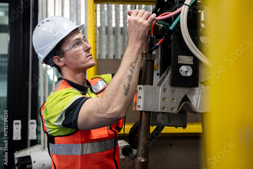 Schilderij op canvas Technician wearing helmet and safety vest performs maintenance on robotic arm industrial machine, symbolizing safety, industry, and engineering skills