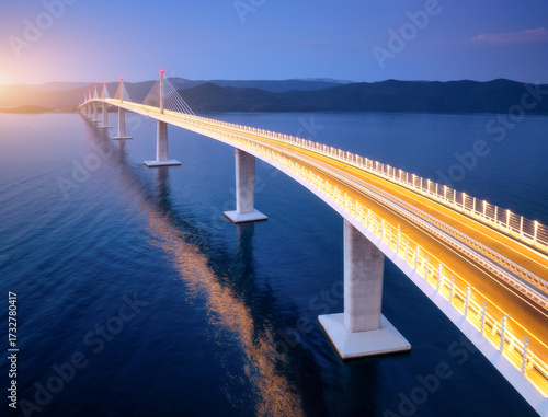 Aerial view of modern Peljesac bridge and blue sea at sunset. Croatia. Bridge with illumination, mountains, reflection and purple sky at sunset. Top drone panoramic view of road, street lights at dusk