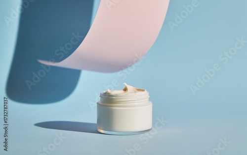 Cosmetic cream jar with pastel abstract shapes casting soft shadows on a blue background
