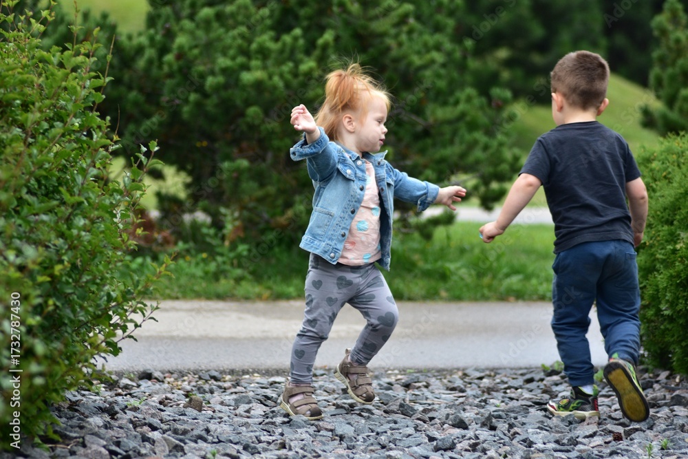 Fototapeta premium Toddler girl in denim jacket and boy in black shirt running happily on gravel path in park. Children enjoying outdoor play, carefree fun, and active childhood friendship moments.