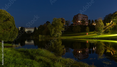 Scenic night view of a riverside park in Narva, Estonia, with modern residential buildings and greenery reflected in the water.
