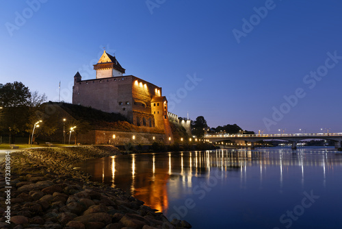 Narva Castle in Estonia illuminated at twilight with reflections in the Narva River, facing Ivangorod Fortress in Russia.