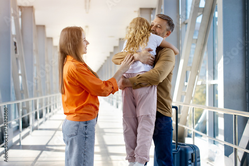 Happy father lifting daughter in arms at airport after arrival, mother standing beside smiling, joyful family bonding moment showing affection, lifestyle and holiday happiness