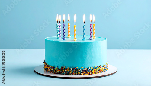 Blue frosted birthday cake with eight lit candles in pink and blue, topped with colorful flowers and berries, set on a white board against a light blue background.